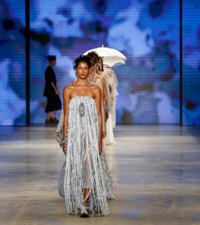 Fila de modelos da Catarina Mina na SPFW N61, com vestido prateado de longas franjas em primeiro plano e sombrinha artesanal ao fundo.